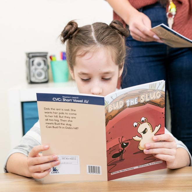 a school-aged girl reading a book in classroom under teacher supervision